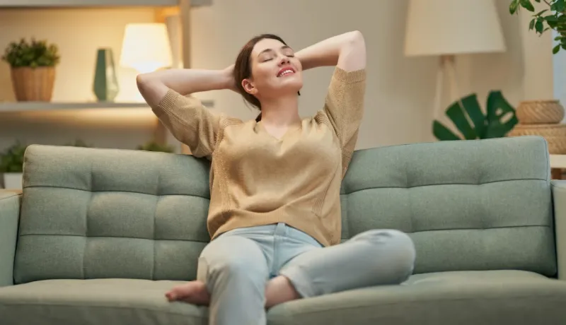 Femme souriante, les bras croisés derrière la tête, se détendant confortablement sur un canapé moderne de couleur verte.