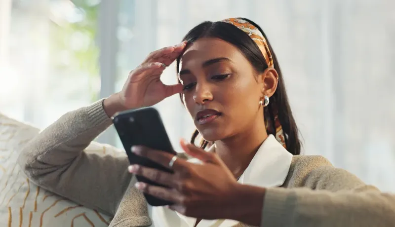 Gros plan d'une femme concentrée ou stressée regardant son smartphone, la main sur son front, dans un environnement lumineux.