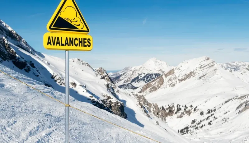 Panneau d'avertissement "AVALANCHES" en jaune vif planté dans une pente de montagne enneigée sous un ciel bleu.