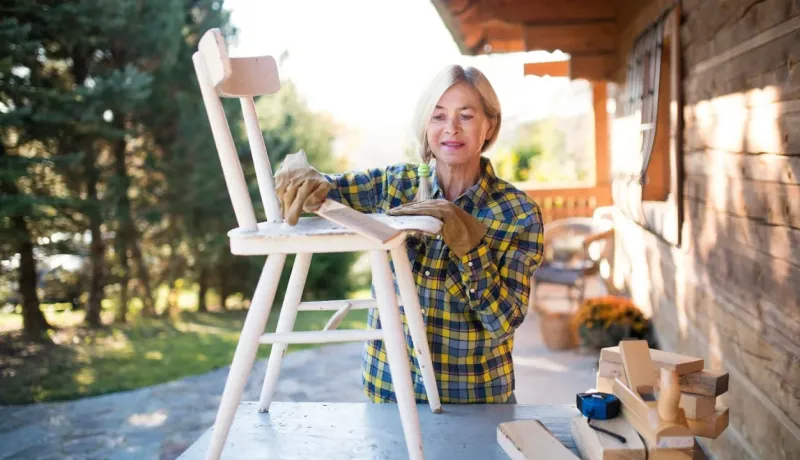 Femme mûre ponçant une chaise en bois sur une terrasse extérieure. Elle porte des gants de travail et une chemise à carreaux.