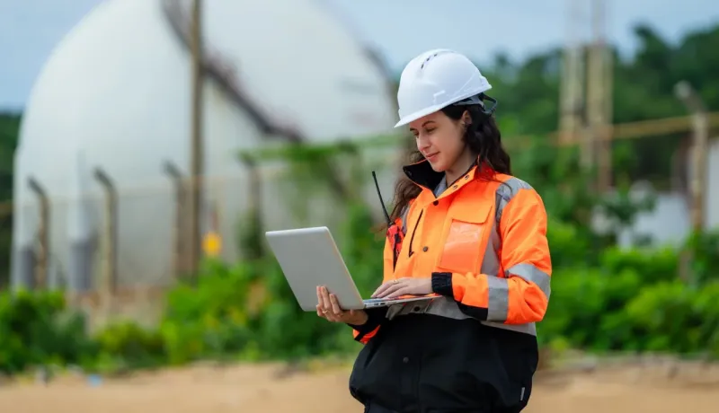 Ingénieure en équipement de protection individuelle (casque et veste orange) utilisant un ordinateur portable sur un site industriel.