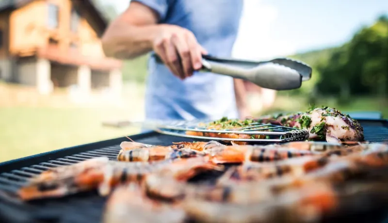 Gros plan sur des crevettes et des légumes grillant sur un barbecue, manipulés par une personne avec une pince.