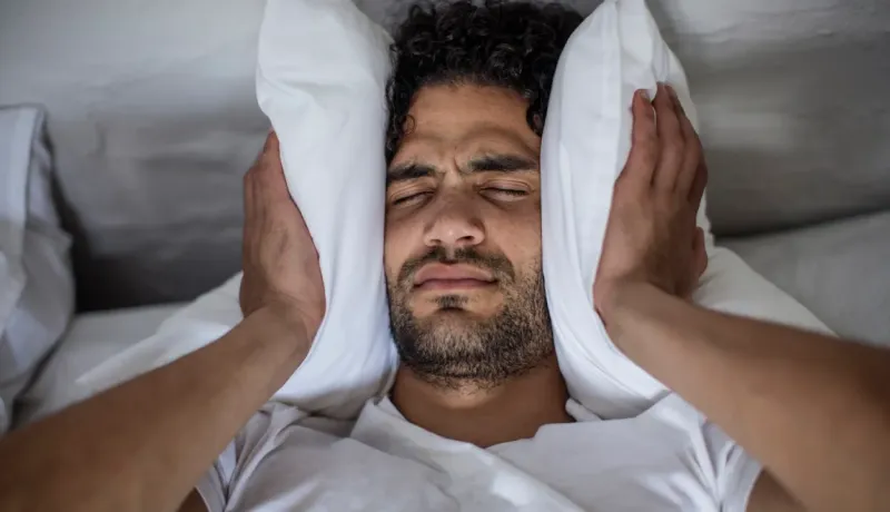 Gros plan sur un homme au lit qui couvre ses oreilles avec des oreillers blancs, l'air irrité par le bruit.