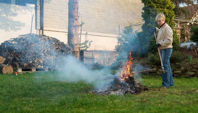 Femme se tenant dans son jardin avec un râteau, regardant un petit feu de branches et de feuilles dégageant de la fumée.