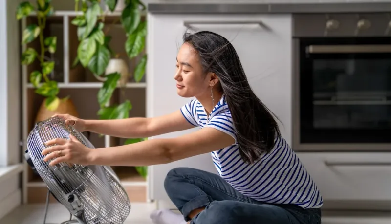 Jeune femme assise sur le sol de sa cuisine, se rafraîchissant devant un ventilateur. Ses cheveux volent légèrement.