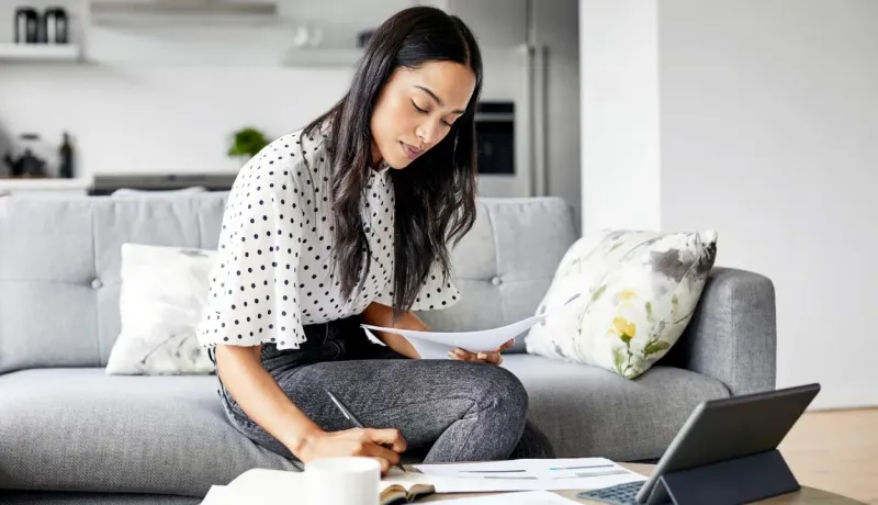 Femme assise sur un canapé, examinant des documents et prenant des notes, travaillant à domicile avec une tablette.