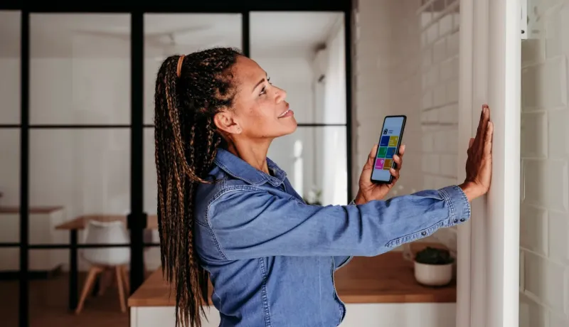 Femme d'âge mûr avec des tresses regardant son smartphone pour contrôler un système domotique mural.