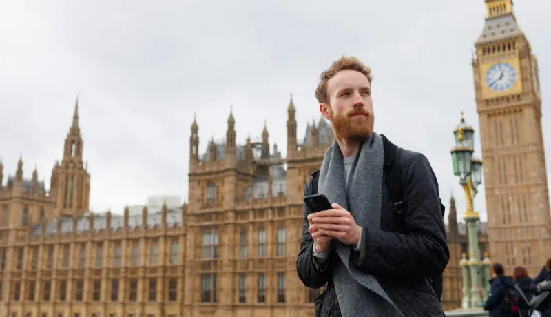 Jeune homme barbu tenant un téléphone devant le Parlement britannique et Big Ben à Londres, par temps couvert.