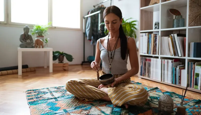 Femme assise en tailleur sur un tapis dans son salon, jouant d'un bol chantant tibétain pour la méditation.