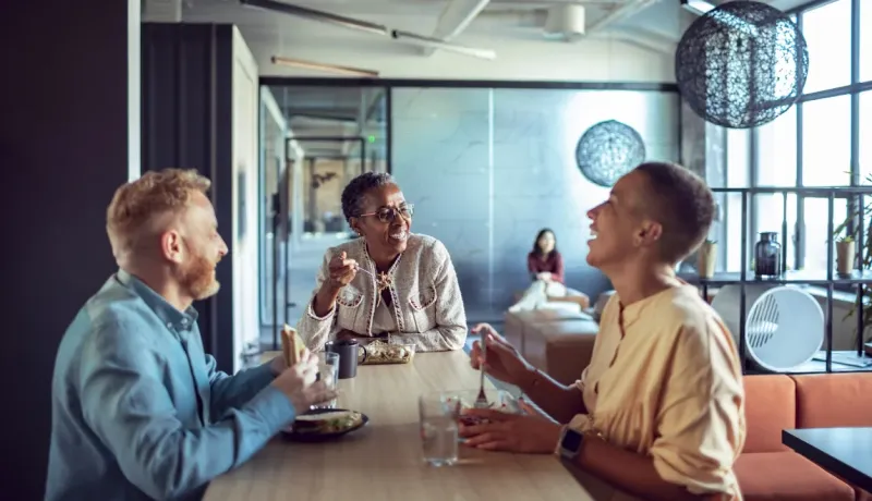Trois collègues de bureau souriants et rieurs prenant leur déjeuner ensemble dans la cafétéria moderne de l'entreprise.