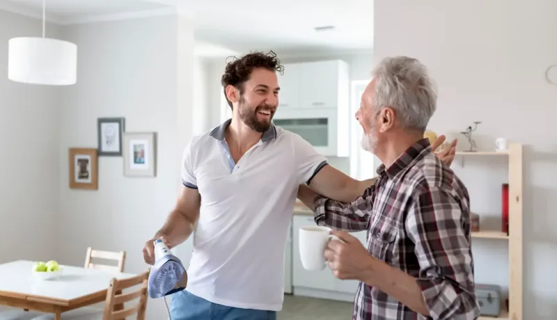 Jeune homme souriant tenant un fer à repasser et un homme plus âgé avec une tasse se saluent affectueusement dans une maison.