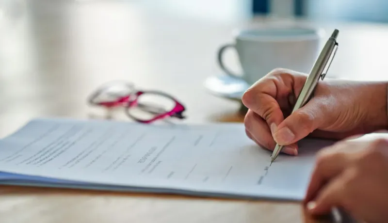 Gros plan sur une main signant un document avec un stylo sur une table, avec des lunettes et une tasse en arrière-plan flou.