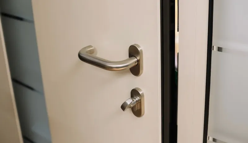 Gros plan sur une porte blanche avec une poignée et une serrure métalliques modernes, un montant vitré est visible à droite.