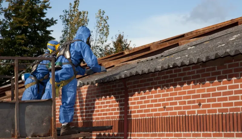 Deux travailleurs en combinaisons bleues de protection et masques retirant une toiture en amiante d'un bâtiment en briques.