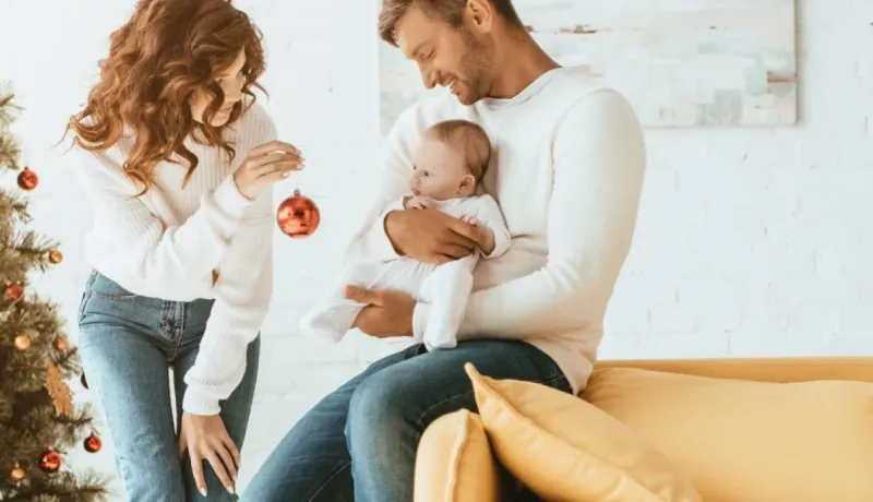 Couple et bébé décorant un sapin de Noël. L'homme tient l'enfant, la femme une boule rouge. Ambiance chaleureuse.