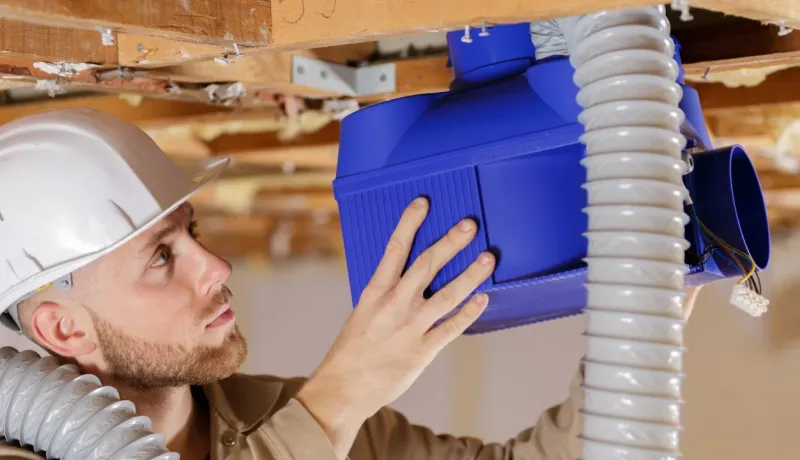 Ouvrier en casque de sécurité installant un système de ventilation (boîtier bleu et gaines) au plafond d'une structure en bois.