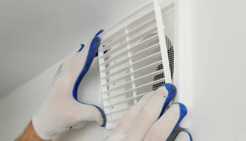 Gros plan sur des mains gantées (blanc et bleu) installant ou ajustant une grille de ventilation blanche sur un mur.
