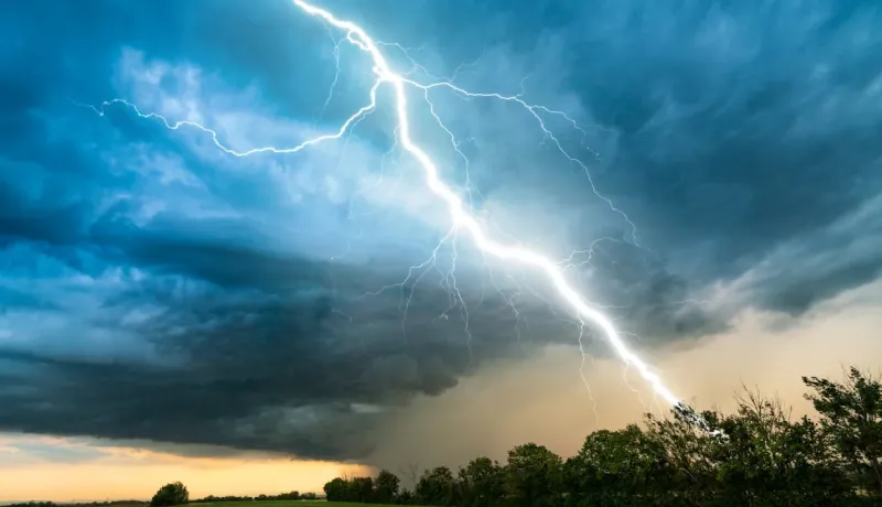 Éclair massif traversant un ciel d'orage bleu foncé et jaune au-dessus d'une ligne d'arbres à l'horizon.