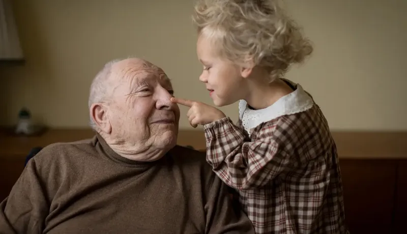 Une petite fille touche le nez de son grand-père avec un sourire, créant un moment tendre et joyeux entre eux.