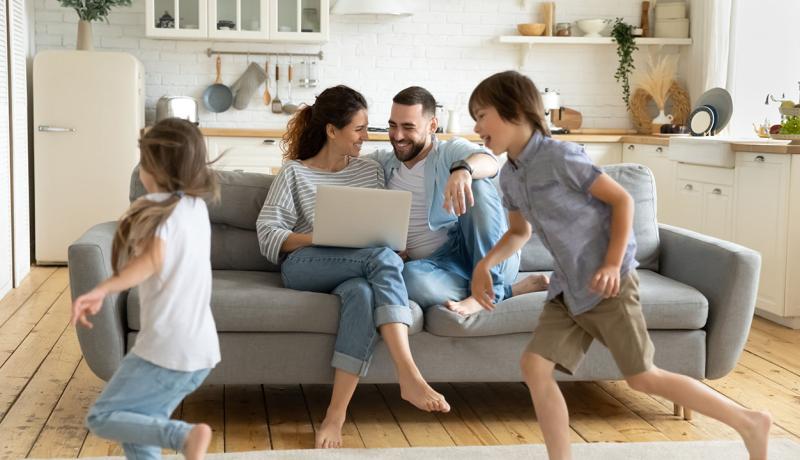 Dans un salon, deux enfants jouent, les parents sur le canapé sont souriants et tiennent un ordinateur sur leur genoux.
