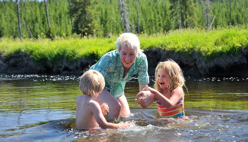 Deux enfants s'amusent dans l'eau, surveillés par leur mère et entourés d'un paysage vert.