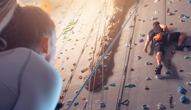 Un homme grimpe un mur d'escalade pendant qu'une femme l'assure depuis le sol.