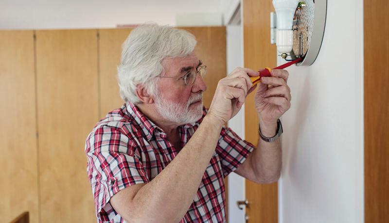 Un homme âgé répare une applique murale avec un tournevis à son domicile.