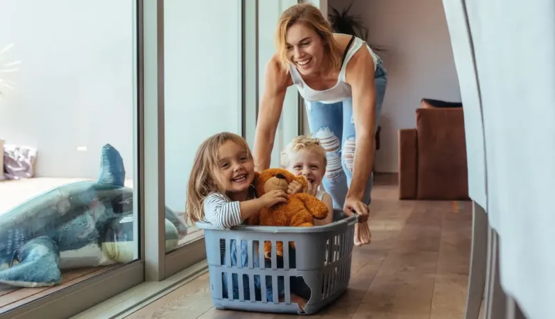 Une mère s'amuse à pousser ses deux jeunes enfants installés dans un panier à linge au milieu du salon.