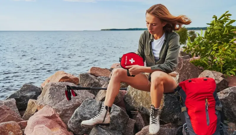 Une femme en randonnée fouille l'intérieur de sa trousse de secours rouge pendant une pause sur des rochers.