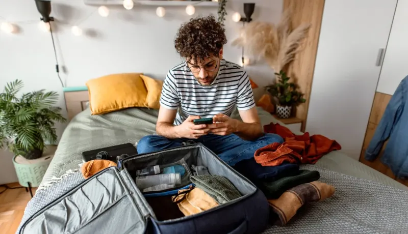 Jeune homme bouclé, valise ouverte devant lui, vérifiant les informations de voyage ou la météo sur son téléphone.