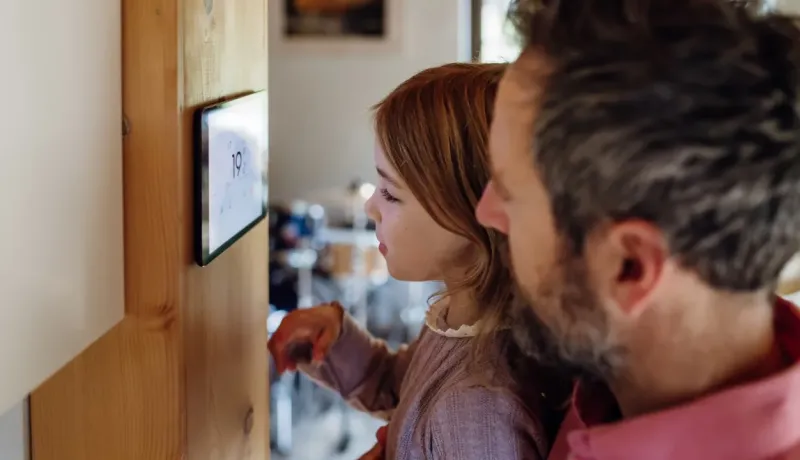 Tête de profil d'une petite fille en pull lilas touchant l'écran d'un thermostat numérique pour régler le chauffage.