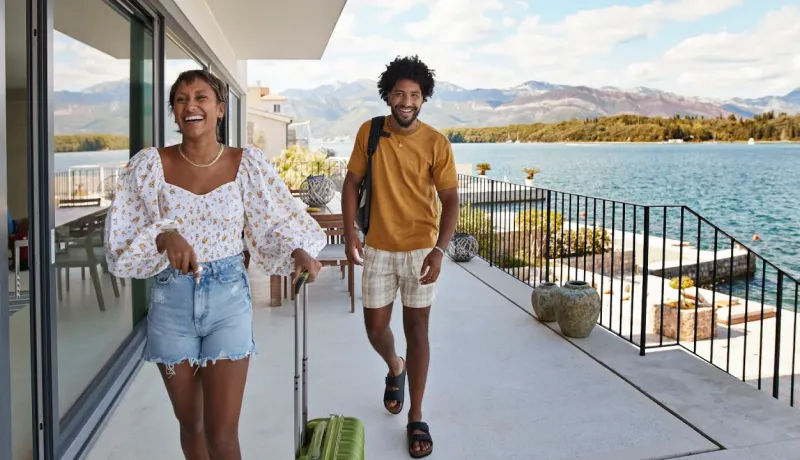 Couple souriant arrivant sur une terrasse de villa avec vue sur la mer et les montagnes. 