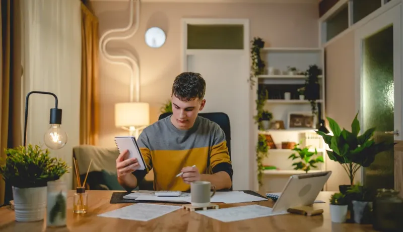 Un jeune homme concentré révise ses finances et prend des notes sur son calepin dans son appartement bien éclairé la nuit.