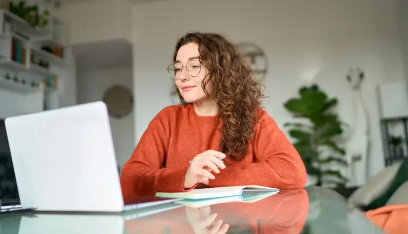 Jeune femme aux cheveux bouclés et lunettes travaille sur son PC portable, prenant des notes.