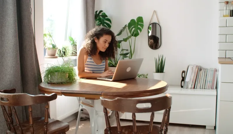 Jeune femme aux cheveux bouclés travaillant sur son ordinateur portable à une table dans une pièce lumineuse remplie de plantes.
