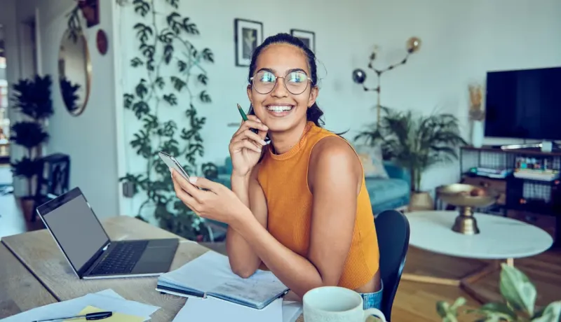 Jeune femme joyeuse et souriante travaillant à domicile, utilisant un smartphone et un ordinateur portable.
