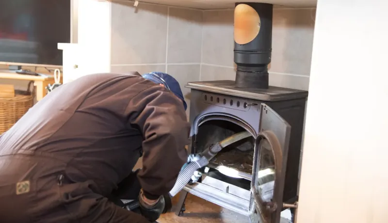 Un professionnel ramoneur nettoie un poêle à bois à l'aide d'un aspirateur dans une maison.