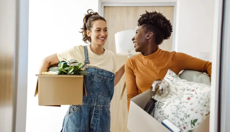 Deux colocataires souriantes déménagent, portant des boîtes remplies d'objets, dont une plante et un oreiller.