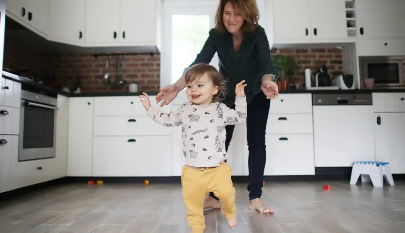 Mère souriante encourageant son jeune enfant à faire ses premiers pas dans une cuisine moderne blanche et brique.