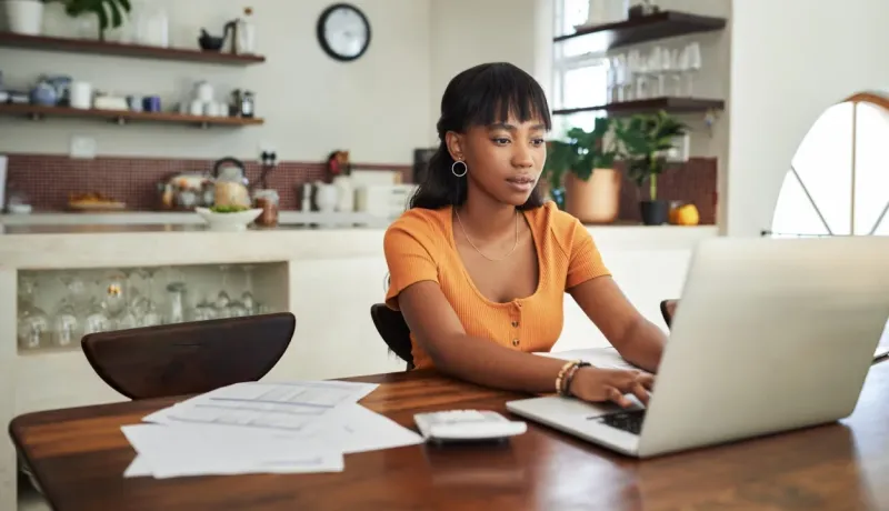 Jeune femme travaillant sur un ordinateur portable à une table de cuisine avec des papiers et une calculatrice.