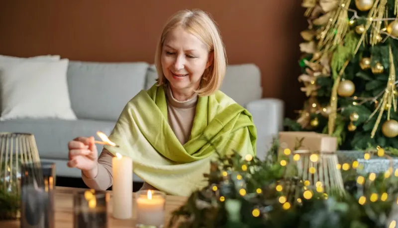 Femme allumant une bougie sur une table décorée pour Noël. Un sapin et des lumières sont en arrière-plan.