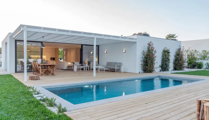Villa moderne avec piscine, terrasse en bois, salon de jardin extérieur et pergola, sous un ciel clair.