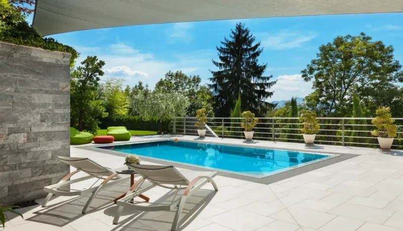 Extérieur luxueux avec piscine, deux chaises longues, terrasse et végétation, sous un voile d'ombrage.
