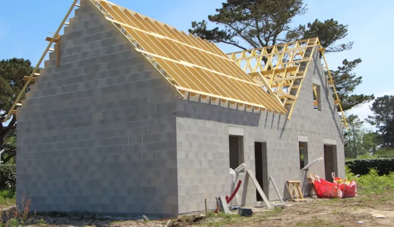 Maison en construction en parpaings de béton avec la charpente de toit en bois exposée.