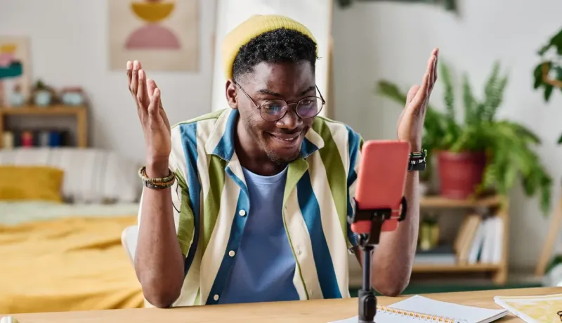 Jeune homme souriant en tenue décontractée faisant un appel vidéo ou un live avec son téléphone sur un trépied.