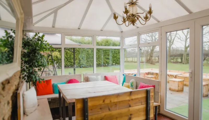 Intérieur d'une véranda lumineuse avec table et banquettes en bois, coussins colorés et vue sur le jardin.