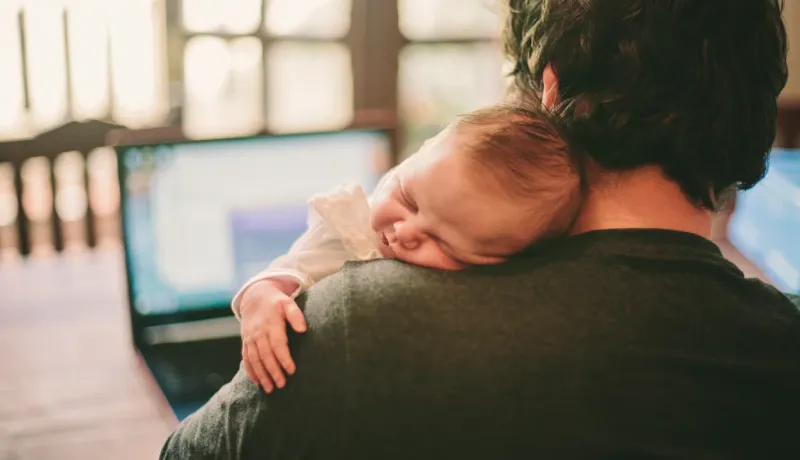 Vue de dos d'une personne devant son ordinateur portable, tenant un bébé endormi sur son épaule dans une ambiance chaleureuse.