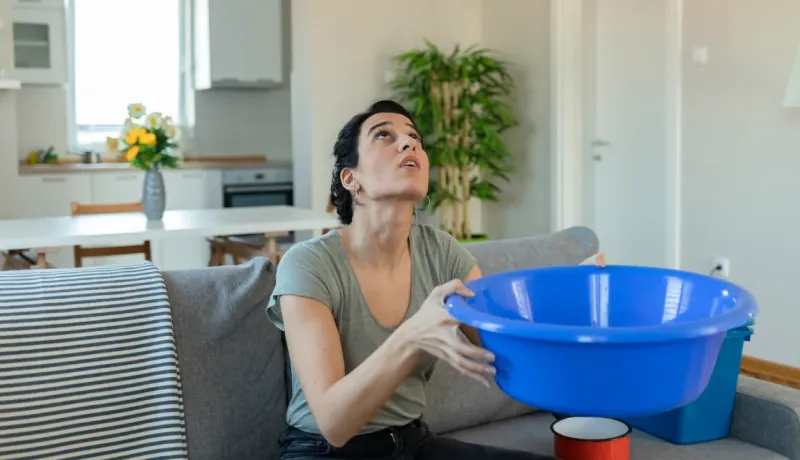 Femme assise sur un canapé, paniquée, tenant une bassine bleue pour récupérer l'eau d'une fuite au plafond.