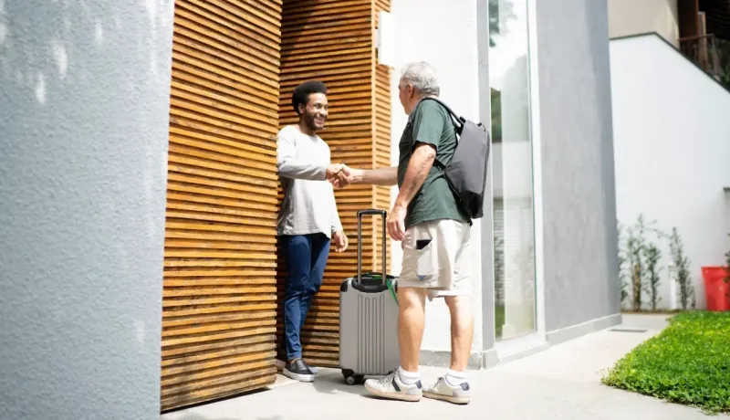Un jeune homme accueillant un homme plus âgé avec une valise et un sac à dos, se serrant la main à l'extérieur.