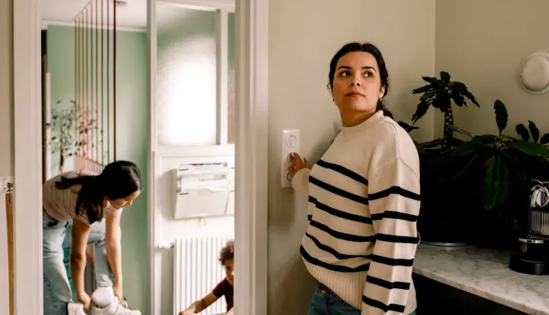 Femme debout appuyant sur un interrupteur ou un thermostat, avec d'autres personnes et enfants en arrière-plan.